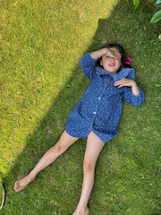 Child lying on green grass in a sunny garden, wearing a blue floral dress, enjoying summer holidays...