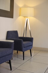 Modern lounge corner with mid-century style blue armchairs, a metal tripod floor lamp, and neutral tile flooring. Minimalist interior with soft ambient lighting and clean, cozy design.