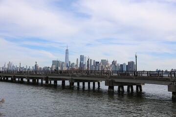 Fototapeta premium Scenic view of the Manhattan skyline featuring the One World Trade Center, seen from the pier in Jersey City across the Hudson River. Urban waterfront cityscape under a partly cloudy sky.