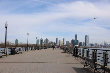 People walking and jogging along a wide waterfront promenade in Jersey City, with a scenic view of...