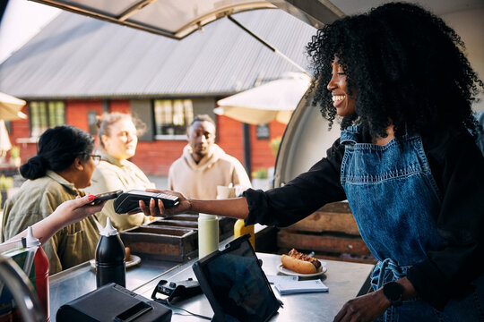 Smiling curly haired female owner giving credit card reader to customer for payment through smart phone in food truck