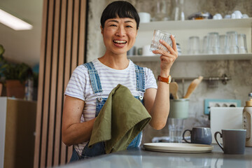 happy mature japanese woman wiping dishes at home kitchen