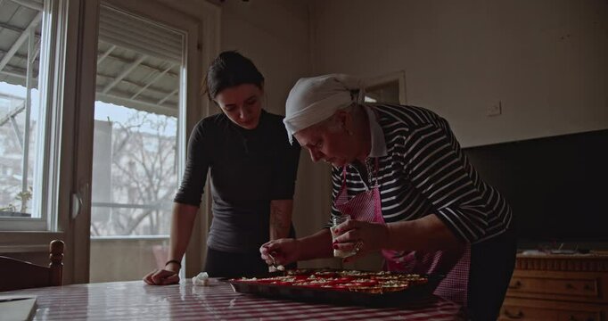 A senior lady carefully prepares a dish, adding a creamy topping to each portion in a baking tray, while a younger woman watches.