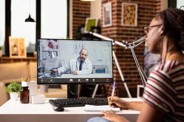 Black female patient talking to caucasian doctor on video call, taking notes during remote medical...