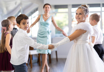 Cheerful junior schoolchildren in party dresses having fun dancing in circle with female pedagogue