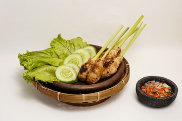 Traditional Balinese satay lilit served on lemongrass sticks with fresh lettuce, cucumber slices, and spicy sambal matah in a stone mortar. Isolated on white background.