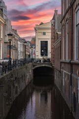 Naklejka premium A canal in Utrecht, Netherlands, with an arched bridge, historic brick buildings, and a white building. Sunset hues reflect on the calm water.