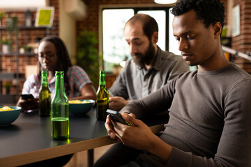 Close up of african american man using smartphone, with bottle of beer nearby on table. Black male individual engages in online browsing while sitting with friends in brick walled room.