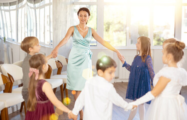 In bright classroom, smiling friendly female teacher joyfully leading circle dance with group of...