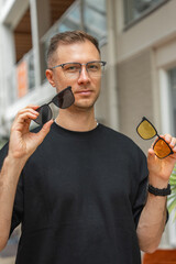 A man with glasses holds sunglasses with dark and yellow lenses indoors. The background shows glass...