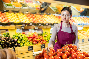 Positive middle-aged female seller putting tomatoes on stand in fruit and vegetable market