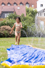 Child joyfully running on a colorful water slide in a sunny garden, surrounded by greenery and...