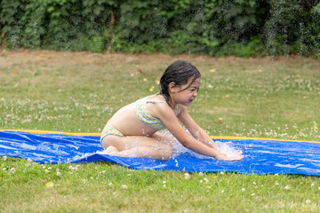 Young girl joyfully sliding on a blue water slide in a sunny garden, splashing water around, capturing the essence of summer fun and playfulness
