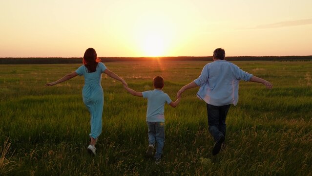 Father mother child son running together holding hands on green meadow at sunset. Family, parents child playing together in sunny park. Slow motion. Picnic dad mom child active recreation in nature