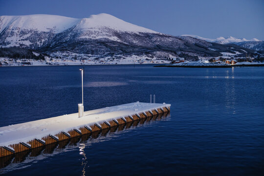 Fototapeta Snow-covered pier on fjord with mountains at twilight