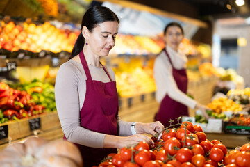Adult woman seller in apron puts tomatoes on display in vegetable shop