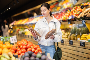 Positive middle-aged woman choosing cassavas standing at counter in large vegetable market