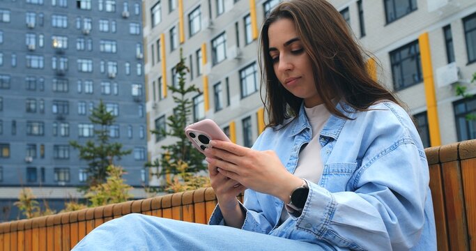 Young woman wearing casual denim jacket sitting on urban bench, browsing internet and engaging with mobile app while enjoying peaceful moment in modern city environment