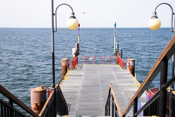 Metal pier with red railings and a lamppost leading to the Baltic Sea on a sunny day. Calm blue water and pale sky in the background. Photo taken in Międzyzdroje, Poland.