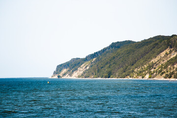 Green hills and sandy slopes with vegetation rise above a blue sea horizon under a clear sky....