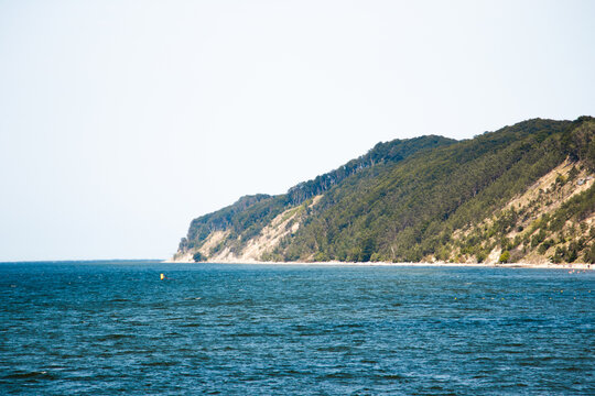 Green hills and sandy slopes with vegetation rise above a blue sea horizon under a clear sky. Coastal landscape with natural textures and contrasting colors.

