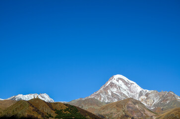 Serene landscape featuring a snow-capped Kazbek mountain, rolling hills with sparse greenery, and a small Gergeti Trinity Church perched atop a distant peak, all beneath a clear blue sky. Georgia