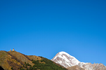Serene landscape featuring a snow-capped Kazbek mountain, rolling hills with sparse greenery, and a small Gergeti Trinity Church perched atop a distant peak, all beneath a clear blue sky. Georgia