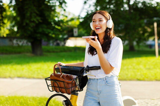 Happy lady near electric bicycle wearing headphones and using cellphone for recording audio message while riding on e-bike in the park