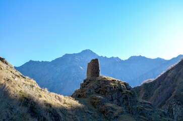 A lone, weathered stone tower stands prominently on a hill, framed by towering mountains and a...