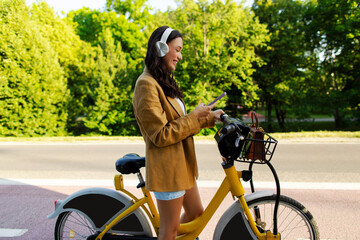 Excited woman with headphones using cellphone, sitting on modern electric bicycle while riding in...