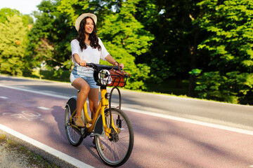 Happy lady in summer outfit and hat riding on renting electric bicycle on bike path in summer sunny...