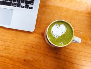 Top view of a green matcha latte with heart-shaped foam art next to a laptop on a wooden table....