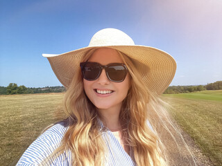 Country path stroll on a bright summer day, woman in wide straw hat and sunglasses smiling beneath...