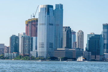 A picturesque cityscape unfolds under a clear blue sky, featuring modern high-rise buildings, waterfront activity, and a sailboat gliding on the water.