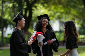 Three graduates in caps and gowns celebrating outdoors. One holds a diploma and bouquet, while the...