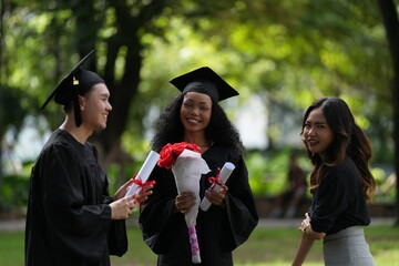 Three graduates in caps and gowns celebrating outdoors. One holds a diploma and bouquet, while the...
