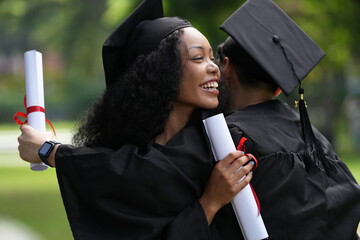 Two graduates in gowns share a warm embrace, holding diplomas with smiles of joy and pride. The...