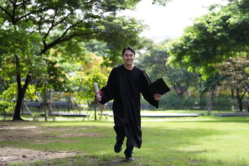 A joyful graduate in a black gown runs across a green field, holding his diploma and a folder,...