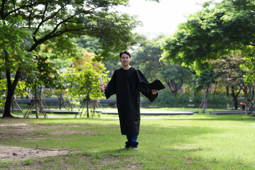 A joyful graduate in a black gown runs across a green field, holding his diploma and a folder,...
