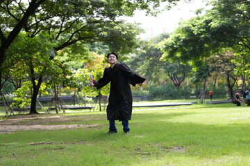 A joyful graduate in a black gown runs across a green field, holding his diploma and a folder,...