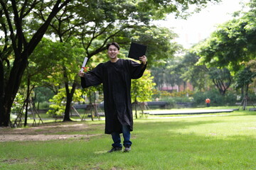 A joyful graduate in a black gown runs across a green field, holding his diploma and a folder,...