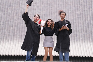 Three graduates celebrate with raised caps and diplomas, smiling joyfully. The two in gowns stand...