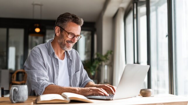 Smiling mature man wearing glasses working on a laptop from a bright modern home office, surrounded by books and natural light, representing productivity, remote work, and digital lifestyle