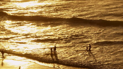 Lisbon, Portugal: Golden waves washing sandy beach, orange sun light reflecting on water, tourists people enjoying sunset over Atlantic Ocean, creating idyllic scene. Summer travel vacation background