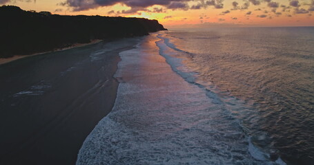 Ocean waves gently rolling onto a dark sand beach at sunset on the coast of Bali, Indonesia, aerial drone footage capturing the beautiful colors of the sky and the peaceful atmosphere