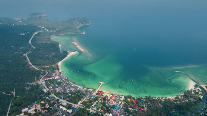 Aerial view capturing a coastal road winding through lush greenery, leading to a vibrant town beside a picturesque bay with crystal-clear turquoise waters on Koh Kood island