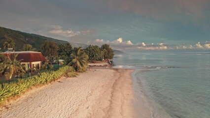 Drone capturing a stunning sunset over a tropical Tahitian beach, with swaying palm trees and traditional houses lining the shore, creating an idyllic paradise