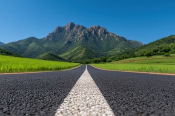 Naklejka premium Low angle view of a long asphalt road with white dividing lines leading to a mountain landscape, green meadow and blue sky
