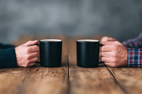 Close up of two people holding black coffee mugs on a rustic wooden table, enjoying a warm beverage and engaging in conversation