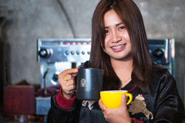 Portrait of a smiling woman holding black and yellow coffee mugs in a cozy cafe. Friendly atmosphere with coffee machine in background. Ideal for lifestyle and beverage themes.